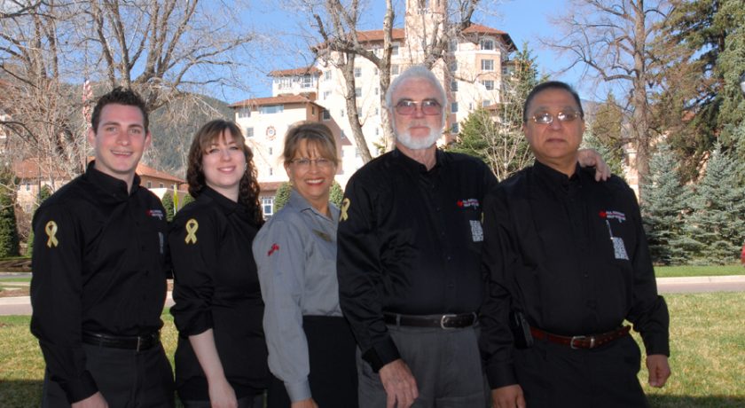 five people wearing grey and black standing, green trees in the background, beige and red building in the middle