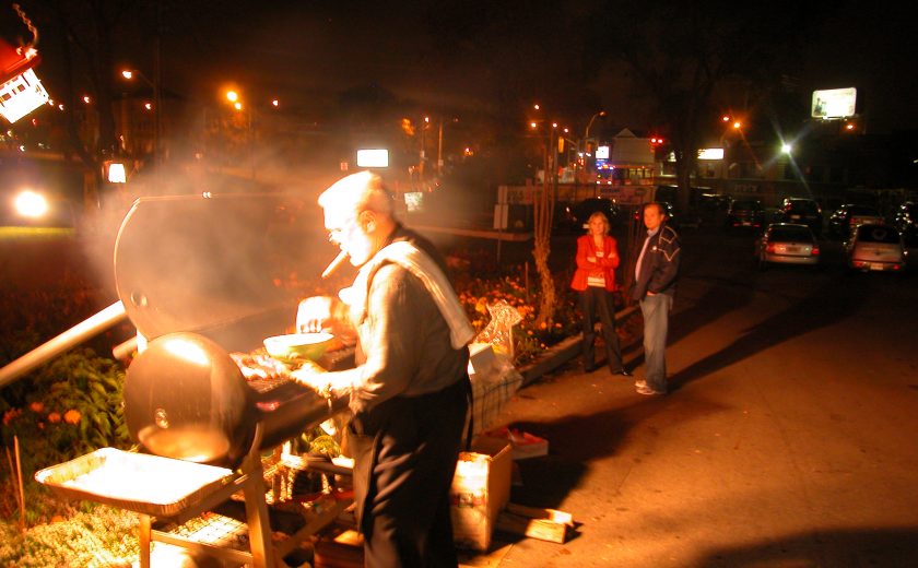 night sky, man barbecuing while holding a bowl, dark filled parking lot