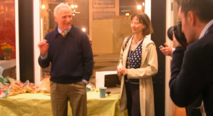 man smiling wearing a dark blue sweater and brown pants, standing in front of table with a yellow table cloth, woman standing with short hair wearing a blue and white polka dot shirt and long beige jacket, man taking picture of man and woman