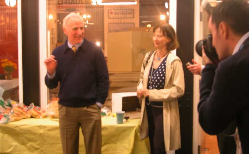 man smiling wearing a dark blue sweater and brown pants, standing in front of table with a yellow table cloth, woman standing with short hair wearing a blue and white polka dot shirt and long beige jacket, man taking picture of man and woman