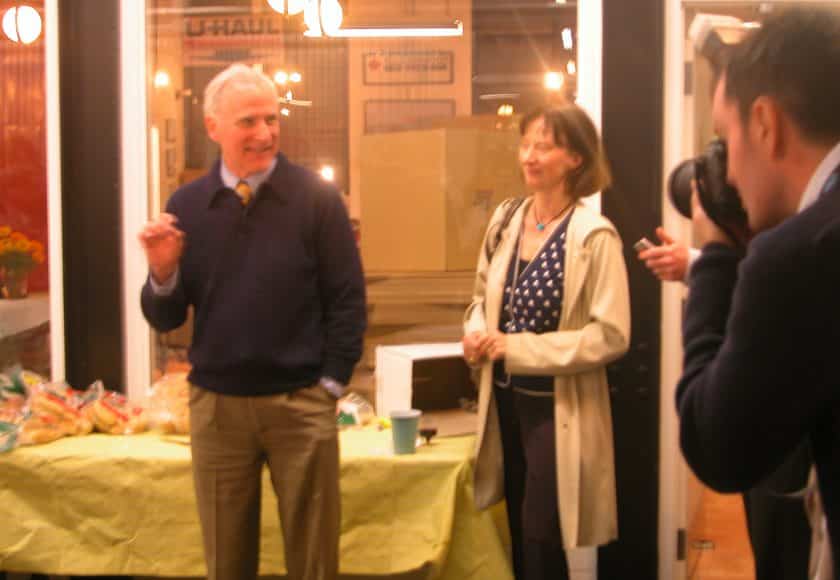 man smiling wearing a dark blue sweater and brown pants, standing in front of table with a yellow table cloth, woman standing with short hair wearing a blue and white polka dot shirt and long beige jacket, man taking picture of man and woman