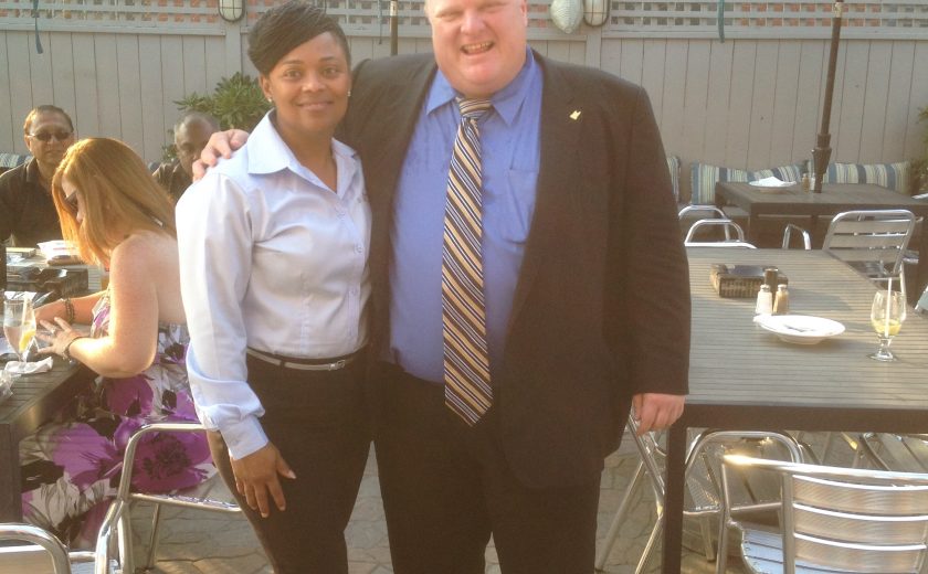 woman wearing a white blouse and black bottoms with black heels, man wearing blue dress shirt and black jacket with striped tie, black tables with silver chairs, teal patio umbrellas