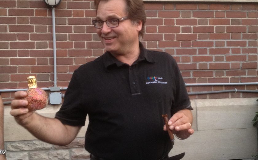 man wearing black shirt holding a cigar and a green circular object, brown brick wall behind, black and white border window