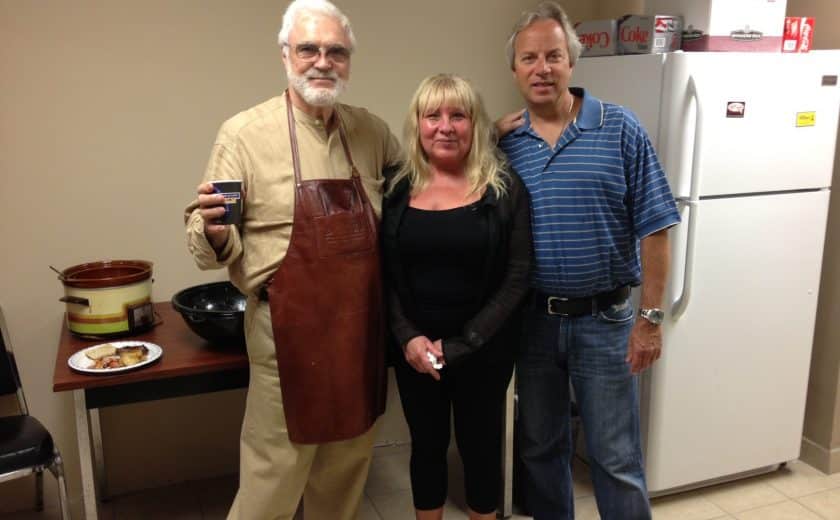 two men and a woman standing in a room with a white fridge and brown table