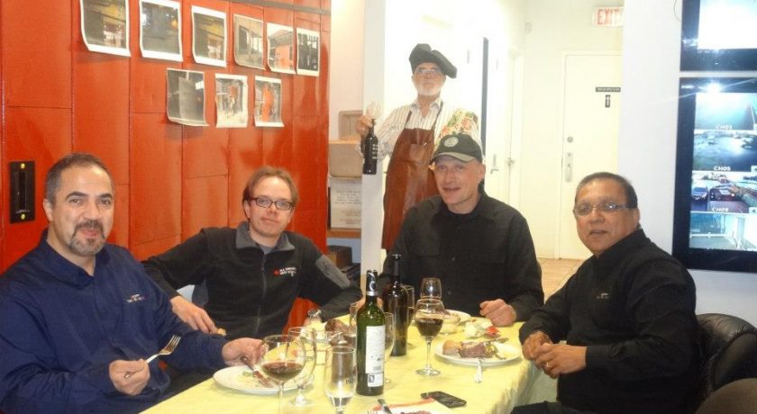 movie night, four men sitting at a rectangular table with a yellow table cloth, wine glasses and alcohol on the table, one man standing in the back holding a black bottle of wine, white and red wall