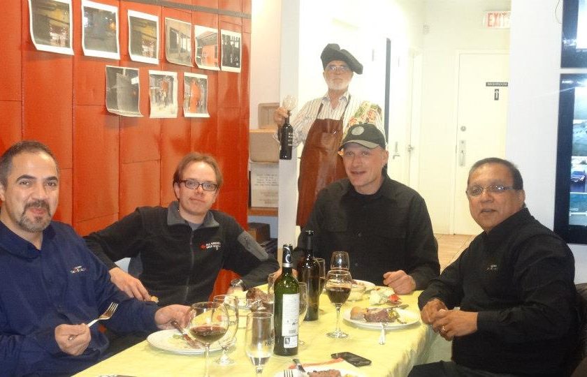 movie night, four men sitting at a rectangular table with a yellow table cloth, wine glasses and alcohol on the table, one man standing in the back holding a black bottle of wine, white and red wall