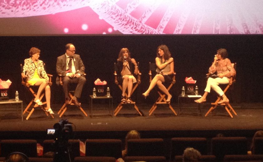 four women and one man on stage sitting on black and brown chairs, pink and black background