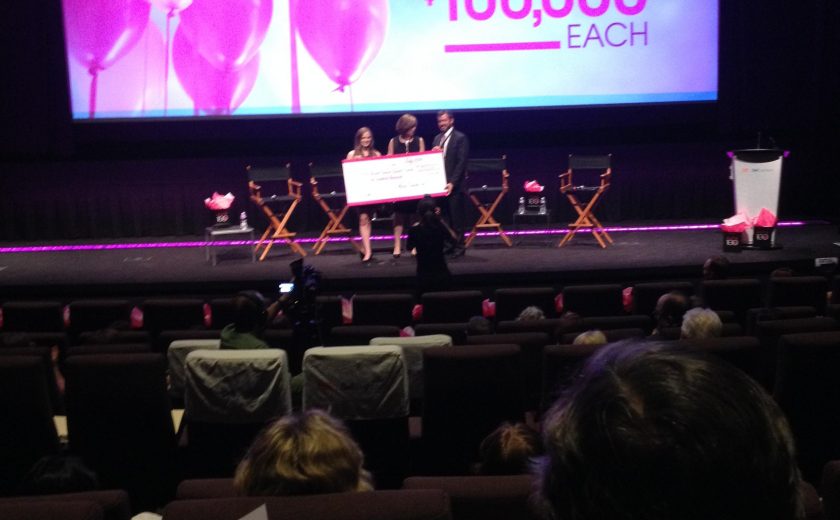 three people on a black stage holding a large cheque, pink and white balloons on the screen