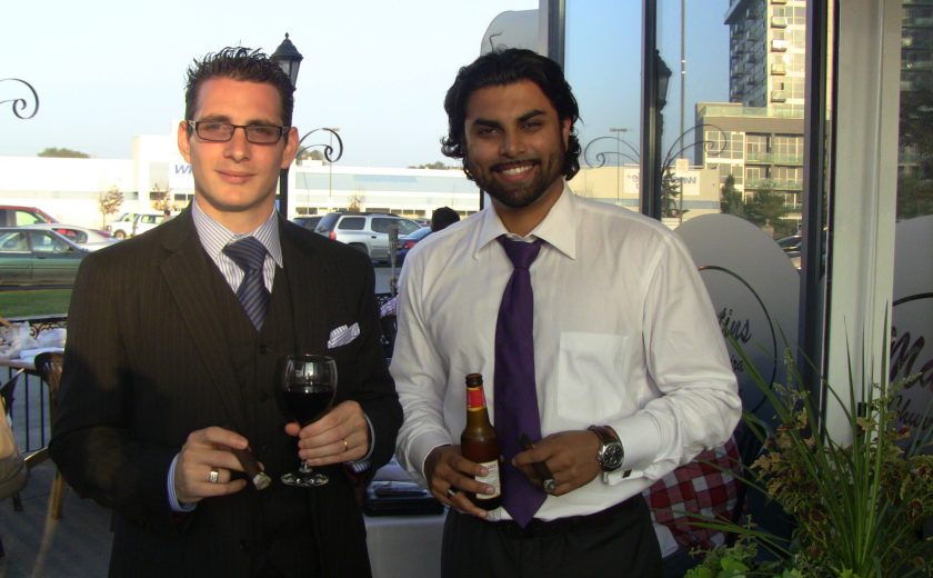 two men smiling, one man wearing a black suit, other man wearing white shirt with purple tie, both holding drinks, blue sky