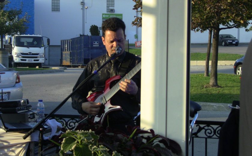 man wearing black, playing a red guitar and singing in a black microphone, white pole on the side, white building in the background