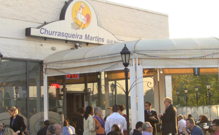 front of Churrasqueira Martins Grill House, white building with white patio tent, people sitting at tables, black gate surrounding