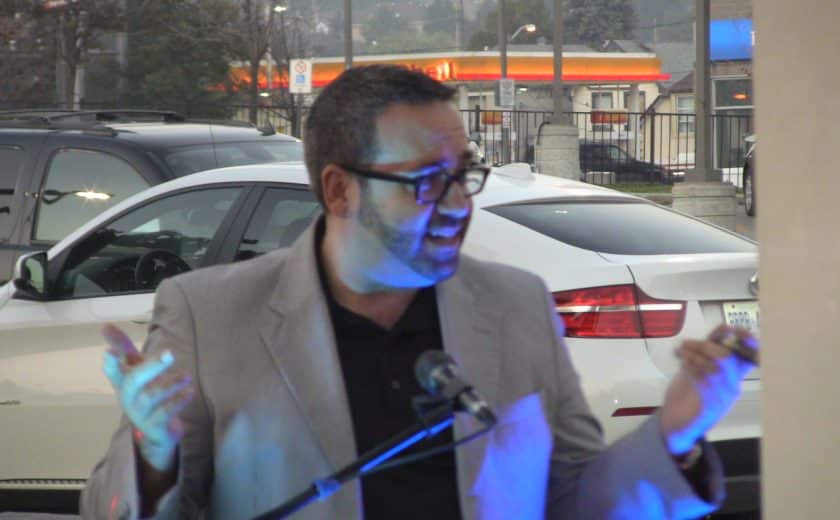 man wearing a black shirt and grey blazer talking on a black microphone, white and black car in the background