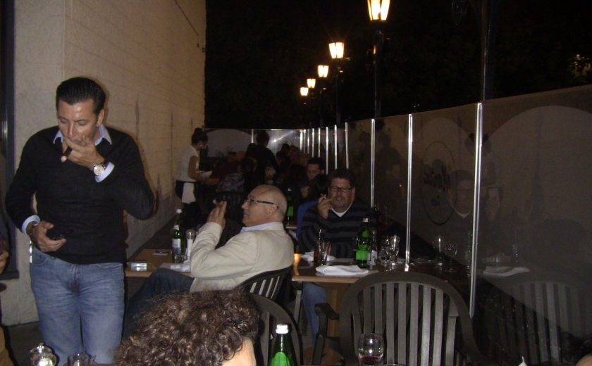 people sitting on black chairs at their brown wooden tables, patio enclosed by glass, 5 tall black lamps in the back, white brick wall on other side of glass boarders