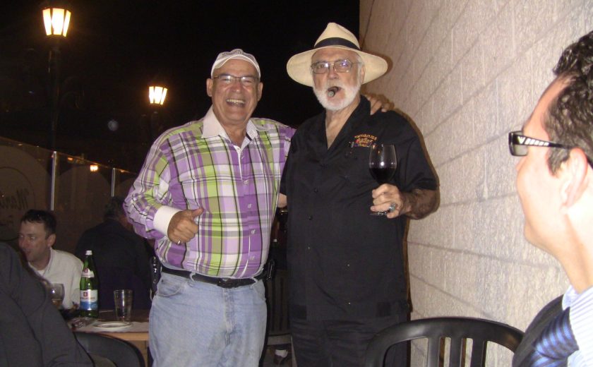 one man wearing purple and green plaid smiling with his arm around another man wearing all black with a cigar in his mouth, night sky, standing beside white brick wall