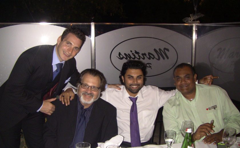 4 men nicely dressed smiling, sitting behind white dinner table, glasses on the table, glass boarder surrounding the patio, night sky