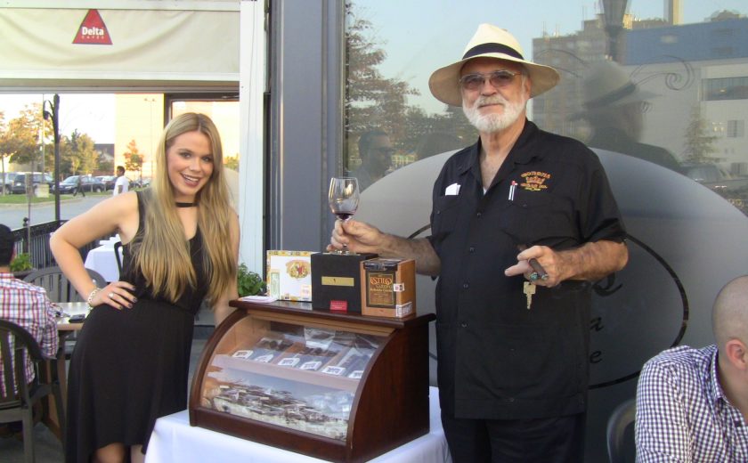 man and woman posing beside mahogany case with glass front full of cigars, case is on white table, outside