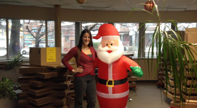 woman wearing red stripes standing beside a red and white inflatable santa, brown boxes stacked around the room, large windows, brown houses outside