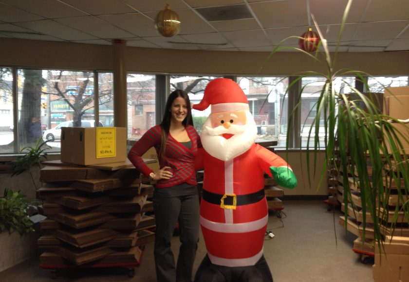 woman wearing red stripes standing beside a red and white inflatable santa, brown boxes stacked around the room, large windows, brown houses outside