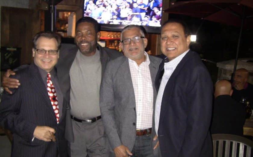 four men standing wearing dress shirts and blazers, tv hanging on the wall behind them