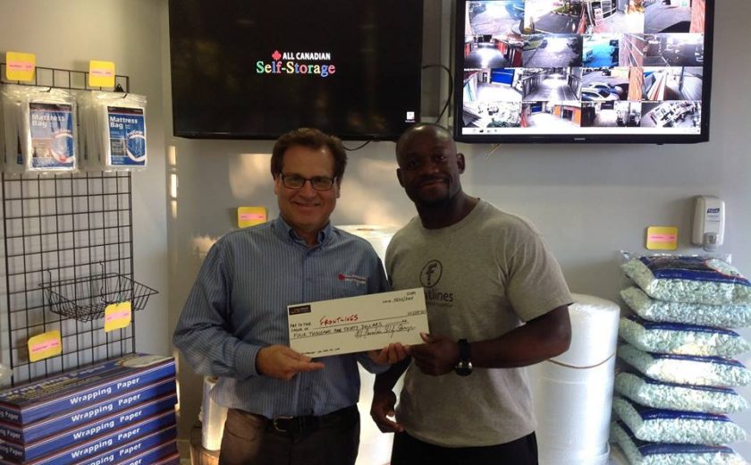 two men standing in front of two screens hanging on the wall, holding a frontlines cheque, surrounded by moving supplies