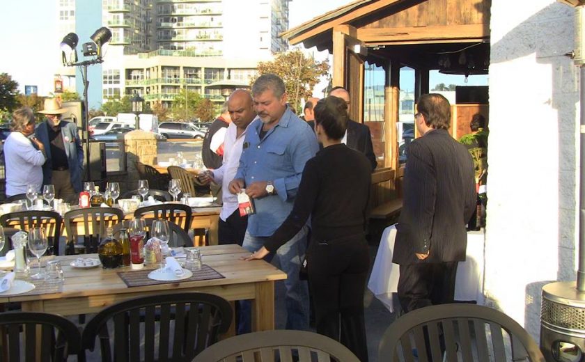 people standing around the wooden tables and black chairs on the patio of the brown and white coloured building, tall buildings and parked cars in the background