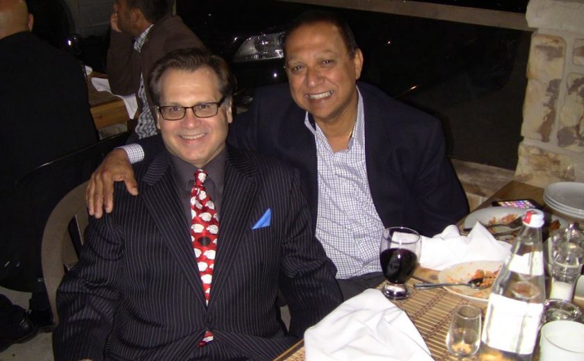 two men wearing dress shirts and jackets, one has their arm around the other, sitting at a dinner table outside with eaten food and utensils on top