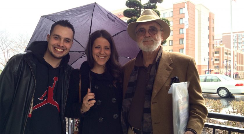 group of three smiling at camera standing, two men on the left and right side, one woman in the middle holding a purple umbrella, tall brown and beige buildings in the background