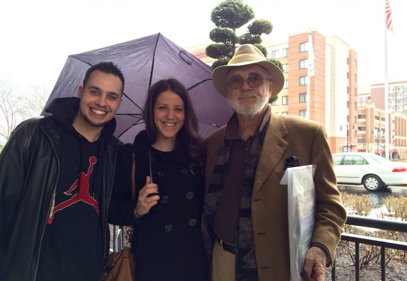 group of three smiling at camera standing, two men on the left and right side, one woman in the middle holding a purple umbrella, tall brown and beige buildings in the background