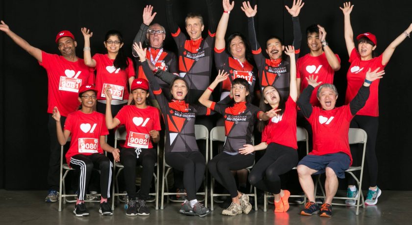 Becel Heart&Stroke Ride for Heart, large group of 15 people smiling and raising their hands up, a mix of wearing red t-shirts and a black and red biking uniform, black background