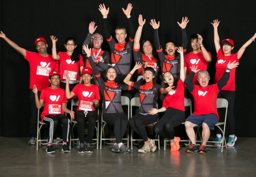 Becel Heart&Stroke Ride for Heart, large group of 15 people smiling and raising their hands up, a mix of wearing red t-shirts and a black and red biking uniform, black background