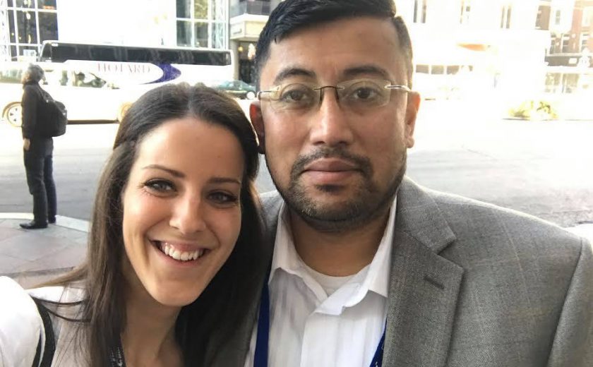 2017 Spring Conference & Trade Show, a man and a woman taking a selfie outside with a building with a lot of windows in the background, both wearing white shirts, man wearing a grey blazer and glasses