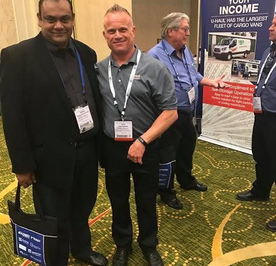 2017 Spring Conference & Trade Show, two men smiling wearing grey shirts and black pants, two other men in the background talking with a U-Haul Increase Your Income backdrop in the background