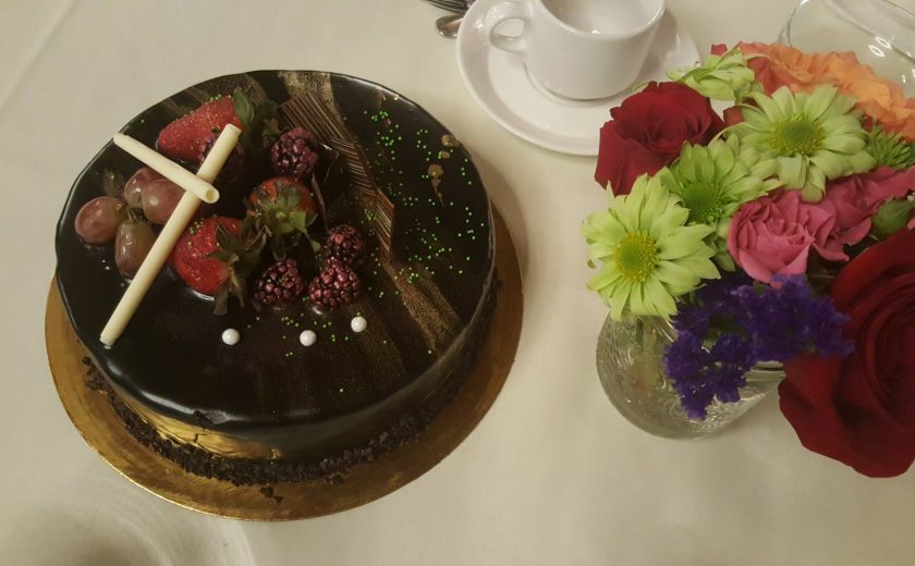 dark chocolate cake with berries and grapes on top, colourful flowers on the right side in a glass jar, white teacup on a white plate behind the cake and flowers