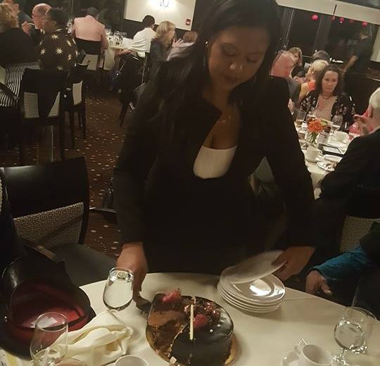 Woman standing passing out plates a round and white dinner table, woman is wearing a white shirt with black jacket, bunch of tables filled with people in the background, bright white lights hanging from the ceiling