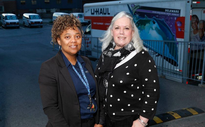 two women smiling, one is wearing a blue shirt with a black jacket, other is wearing a black and white polka dot shirt, U-Haul trucks parked