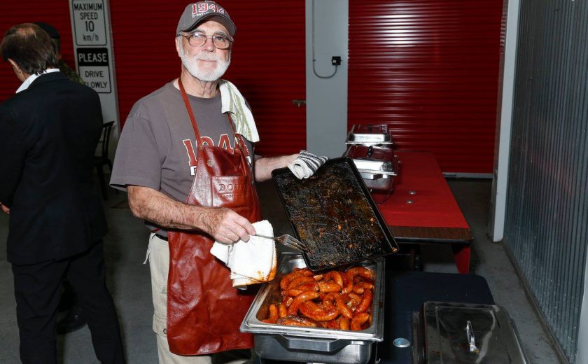 man wearing a grey shirt with red apron, holding a white towel and a black tray, putting red sausages into a silver container, red storage units in the back