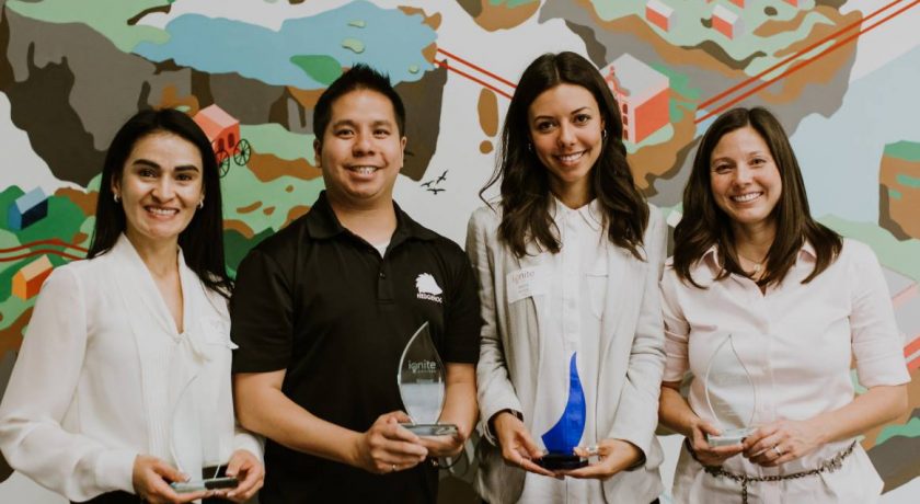 three women and one man standing with clear and blue trophies from ignite capital pitch competition, women are wearing white and pink blouses, man wearing black t-shirt, colourful design on the wall