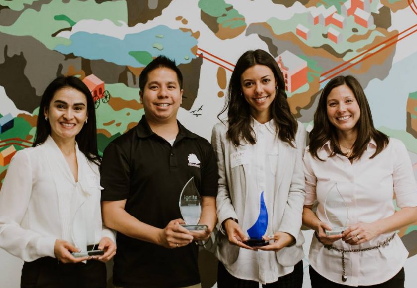 three women and one man standing with clear and blue trophies from ignite capital pitch competition, women are wearing white and pink blouses, man wearing black t-shirt, colourful design on the wall