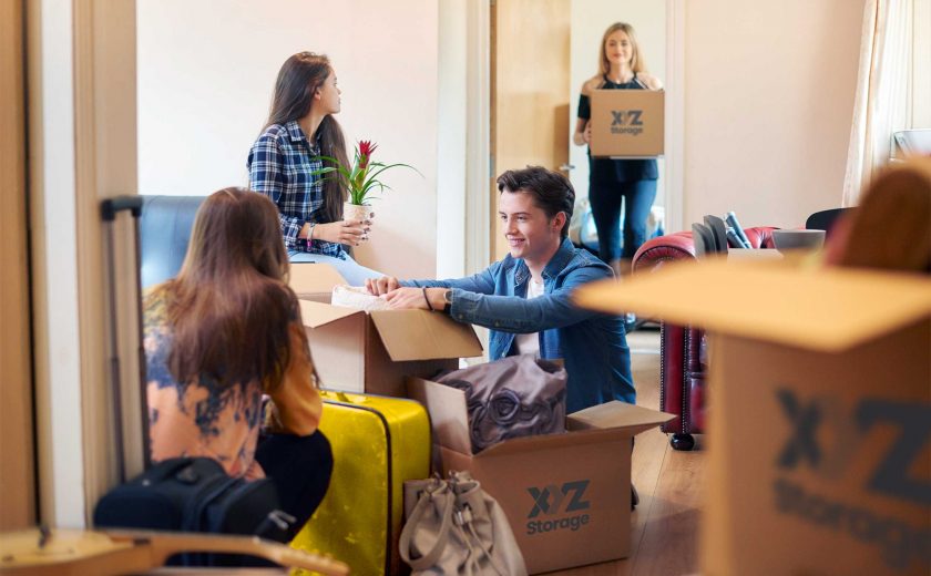 boy and girl on the floor packing their apartment with XYZ Storage moving boxes, one girl sitting on a couch holding a plant, another girl standing holding an XYZ Storage moving box