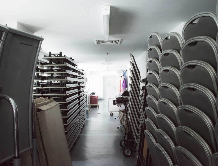 white storage room with multiple black chairs, tables, boxes, and colourful clothes on a hanger in the back