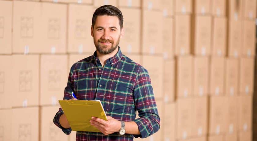 man wearing a colourful plaid shirt holding a yellow clipboard a blue and white pen, wall of brown boxes in the background