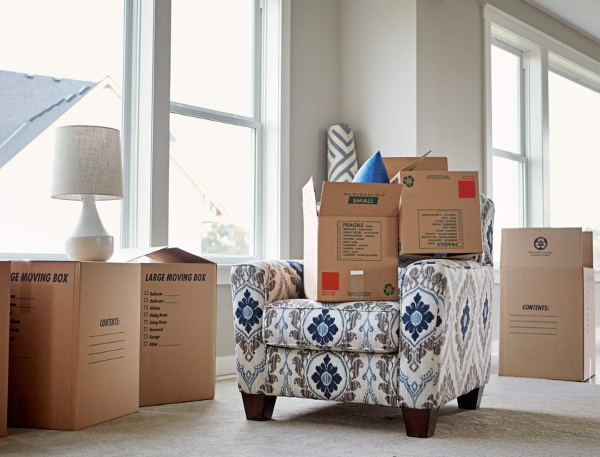 white room with windows and a lot of natural lighting, several brown moving boxes, white armchair in the middle with a blue and grey design