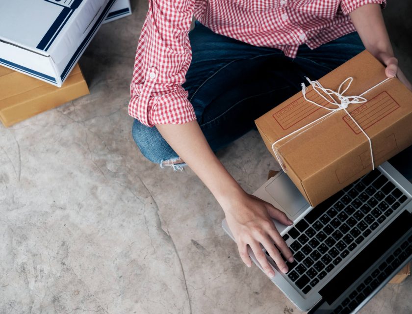 person wearing a red and white plaid shirt and blue jeans, holding a brown package with a white string, typing on a laptop