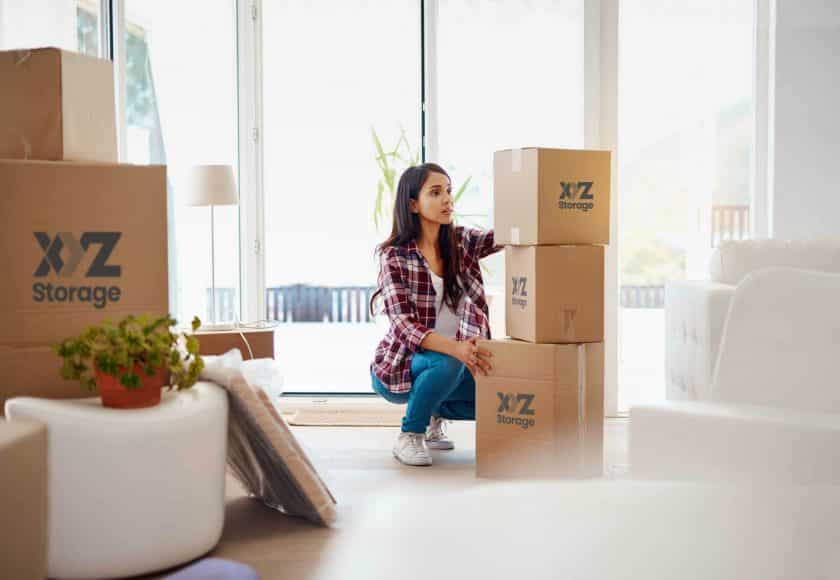 young woman wearing a plaid shirt with blue jeans and white shoes, packing her belongings using XYZ Storage boxes