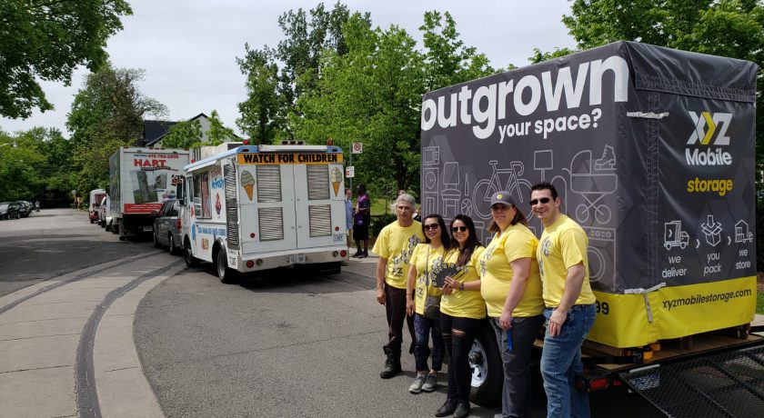 XYZ Storage team wearing grey and yellow standing in front of large grey and yellow mobile storage container, white ice cream truck in front, green trees in the background