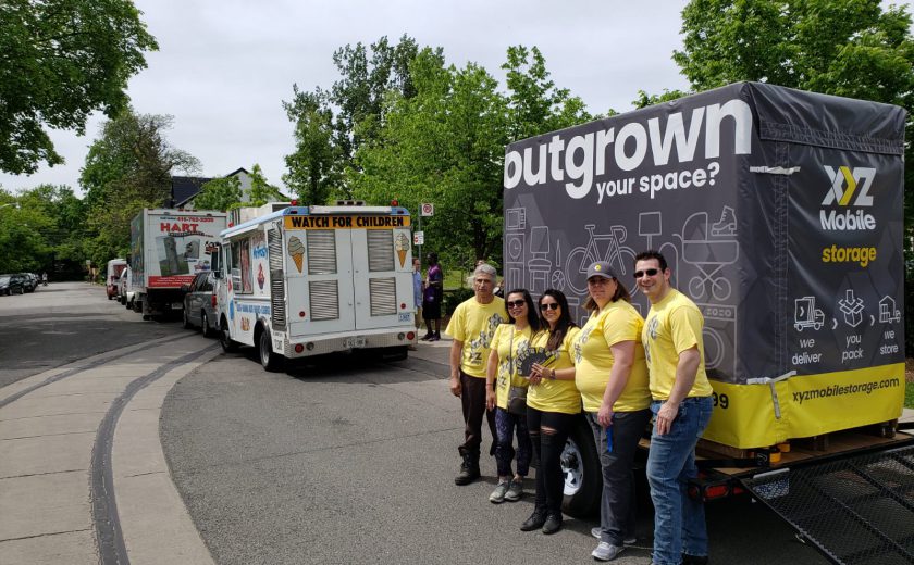 XYZ Storage team wearing grey and yellow standing in front of large grey and yellow mobile storage container, white ice cream truck in front, green trees in the background