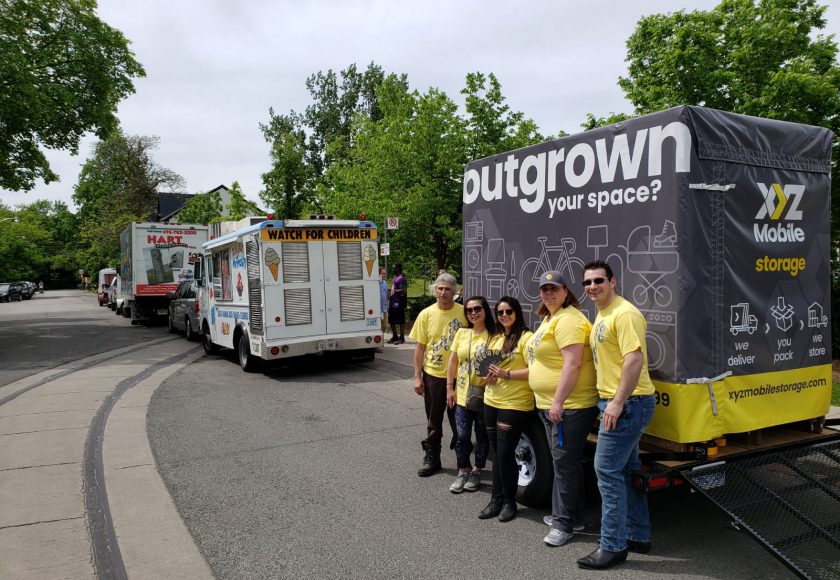 XYZ Storage team wearing grey and yellow standing in front of large grey and yellow mobile storage container, white ice cream truck in front, green trees in the background