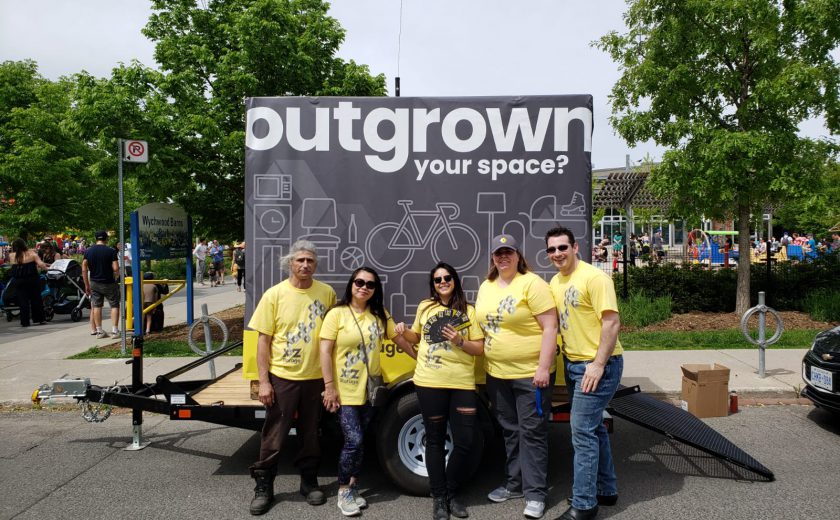 XYZ Storage team wearing yellow and grey standing in front of white and grey mobile storage container, standing in front of a park with children and green trees