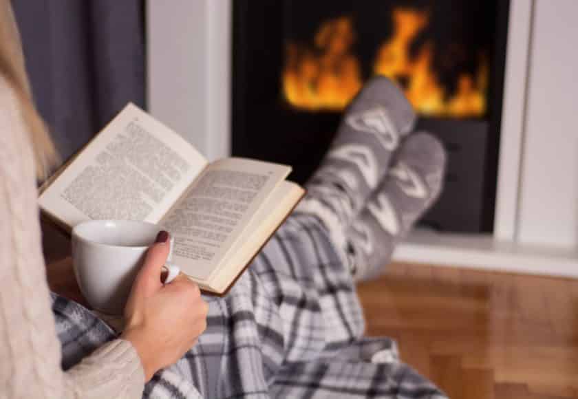 Beautiful young woman in front of the fireplace reading book and warming feet on fire and legs are covered with blanket, in hand holds cup of hot tea. Winter and cold weather concept at home. Close up, selective focus