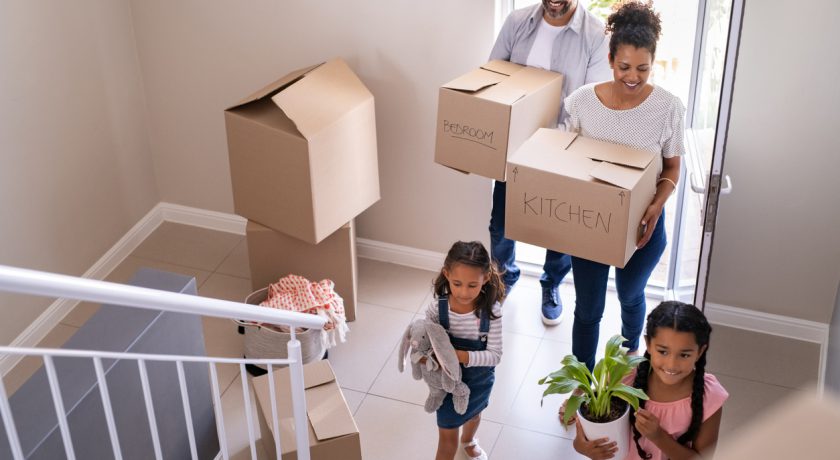 Ethnic family with two children carrying boxes and plant in new home on moving day. High angle view of happy smiling daughters helping mother and father with cardboard boxes in new house. Top view of excited kids having fun walking up stairs running to their rooms while parents holding boxes.